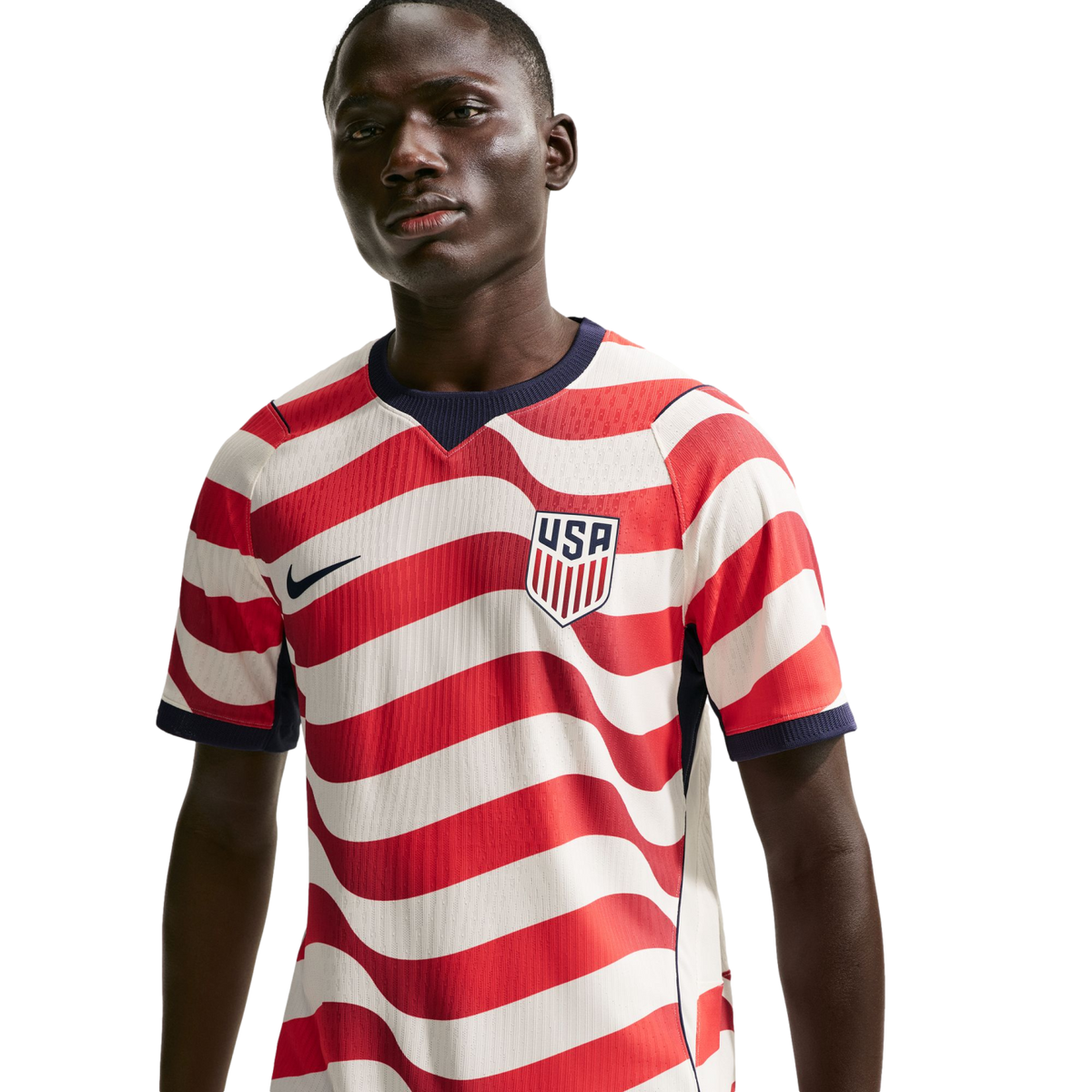 A man wearing a USA soccer jersey with red and white wavy stripes, featuring a Nike swoosh and USA crest, against a solid black background.