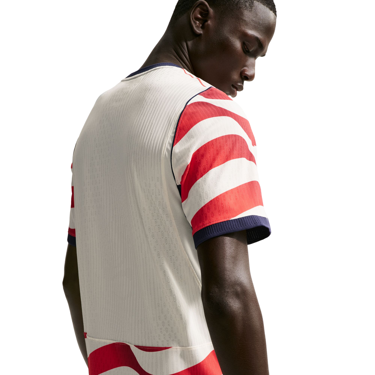 Rear view of a man looking down, wearing a white soccer jersey with red and white striped sleeves and navy blue trim against a black background.