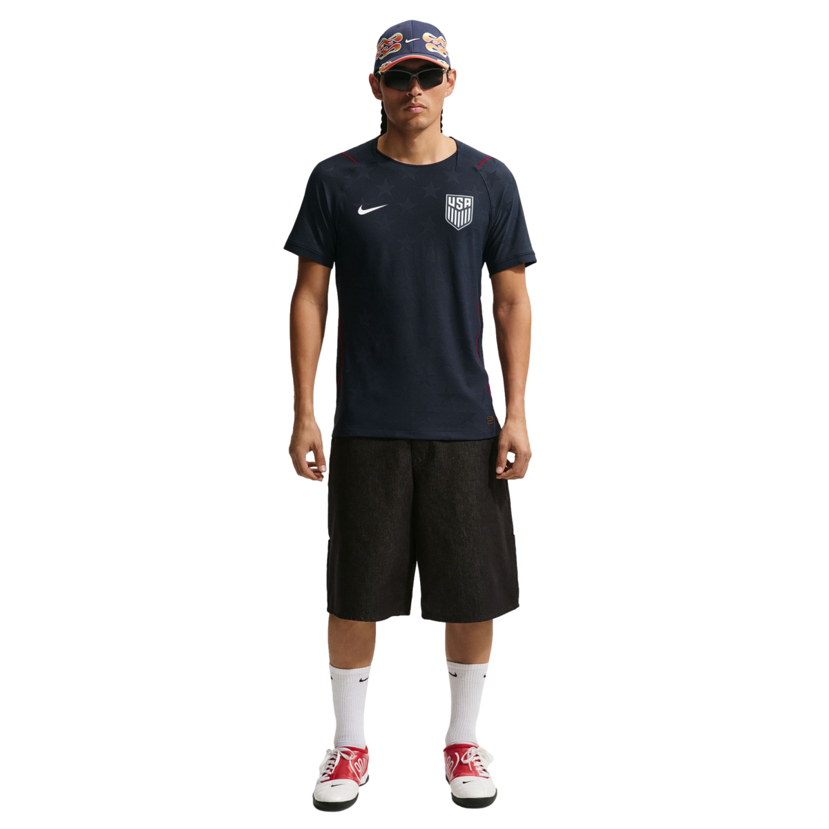 A man in a navy USA soccer jersey, black shorts, and red sneakers stands against a black background wearing a patterned cap and sunglasses.