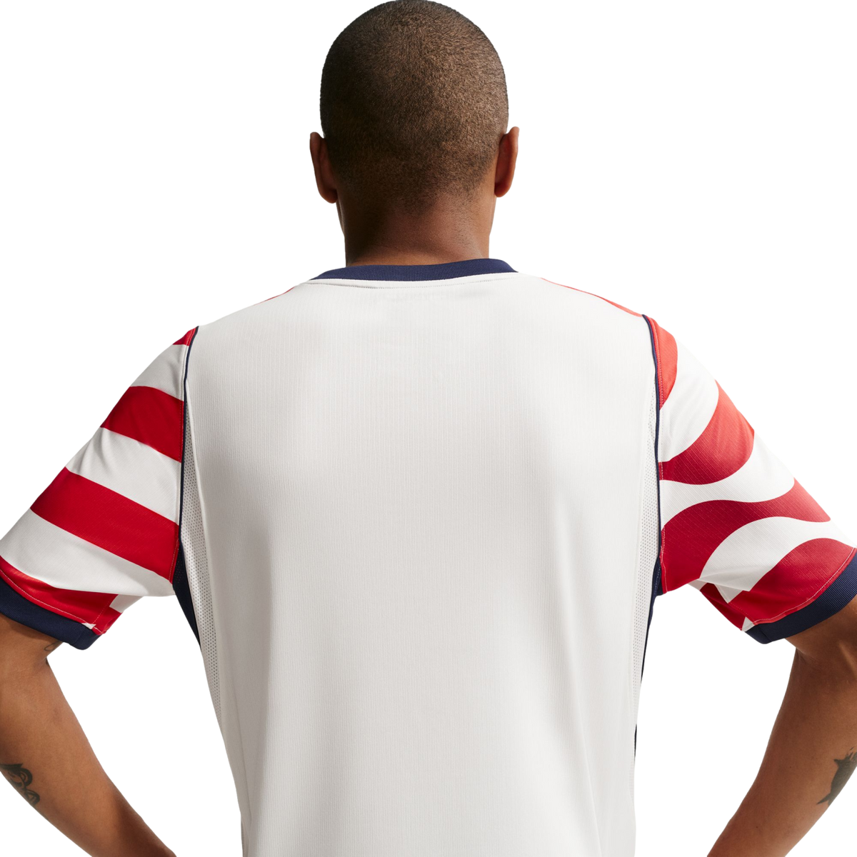 Rear view of a man with a buzz cut wearing a white jersey with red-and-white patterned sleeves and navy trim, standing with hands on hips against a black background.