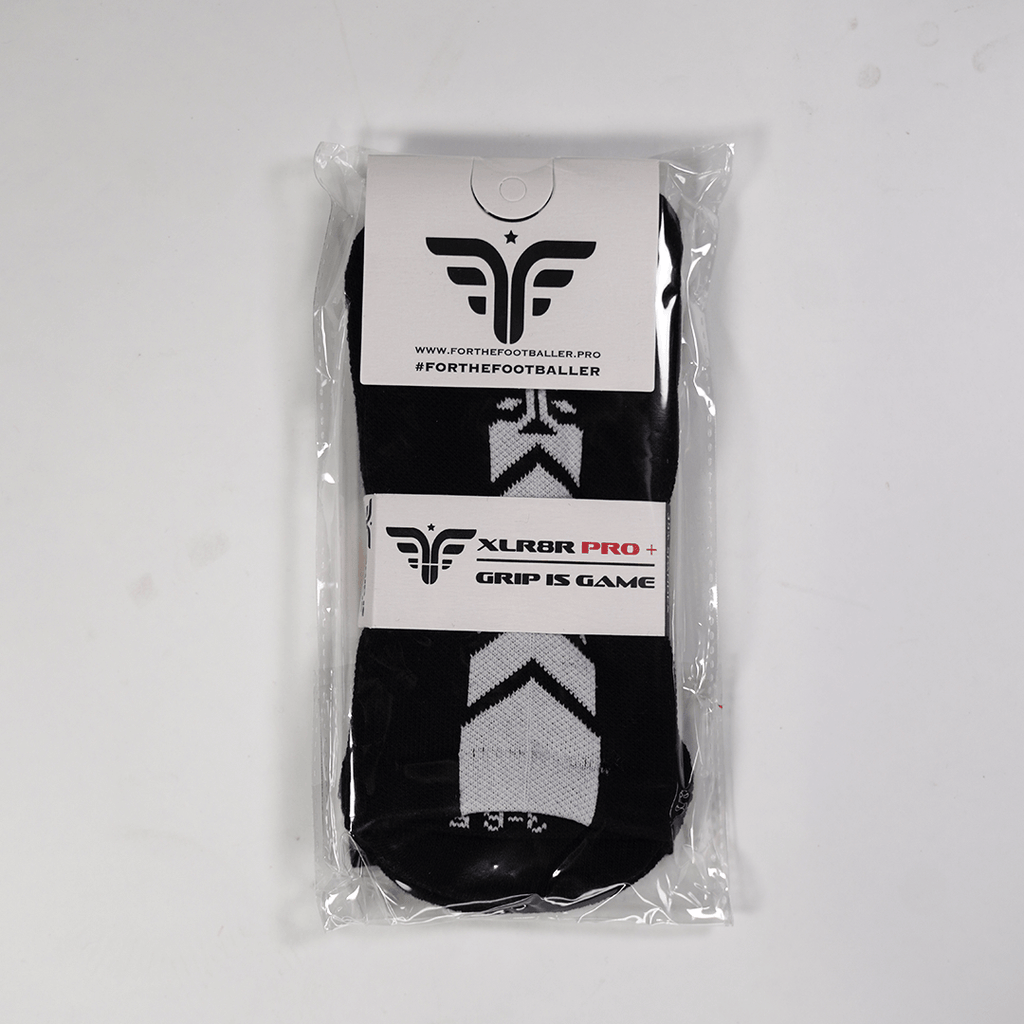 A pair of For The Footballer XLR8R Pro No Slip Socks - Black, shown in clear plastic packaging. The black socks feature white chevron-like patterns and are held by a white cardboard header and a middle band with the brand logo and "XLR8R PRO+" text.