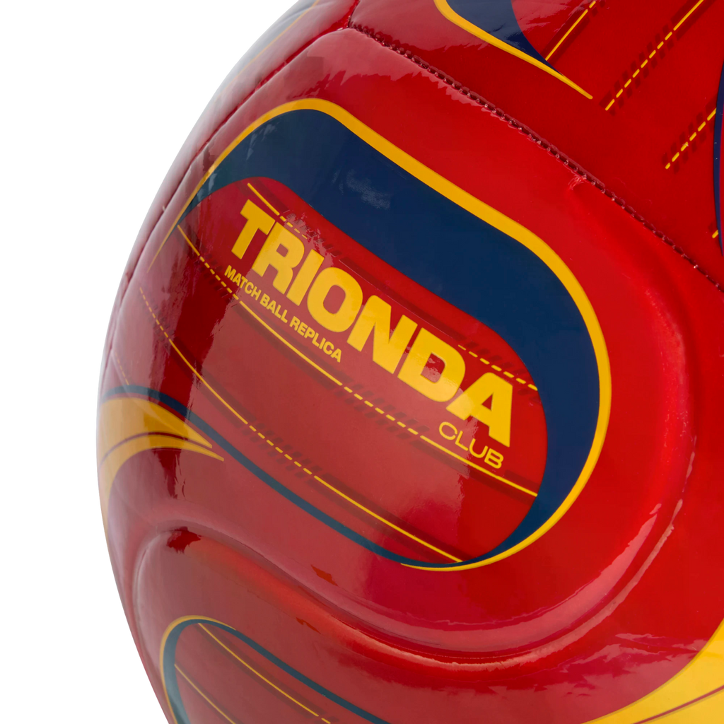 Close-up of a red soccer ball with yellow and blue graphics, featuring the yellow text "TRIONDA CLUB MATCH BALL REPLICA" against a black background.