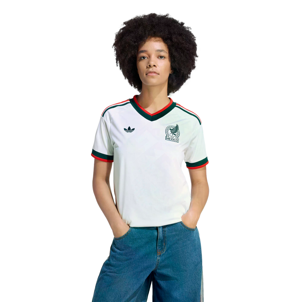 A person with dark curly hair wears a white Mexico Adidas soccer jersey with green and red trim and blue jeans against a solid black background.