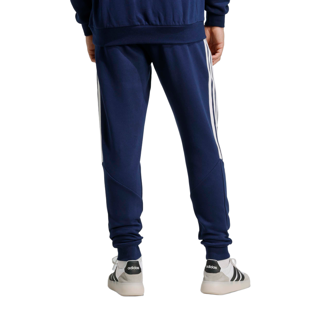Rear view of a person wearing navy Adidas track pants with white side stripes and white sneakers against a light gray background.