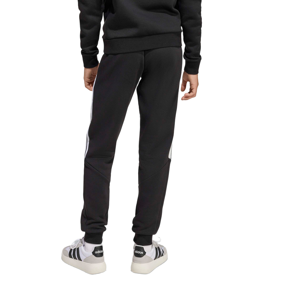 Rear view of a person wearing a black sweatshirt, black Adidas sweatpants with white side stripes, and white Adidas sneakers against a plain light grey background.