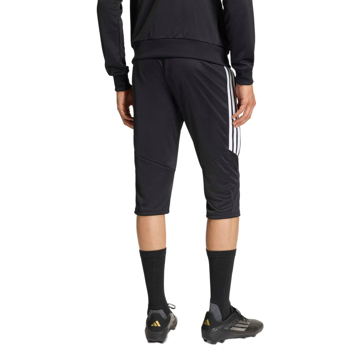 Rear view of an athlete wearing black three-quarter length soccer training pants with white side stripes and black soccer cleats against a plain background.