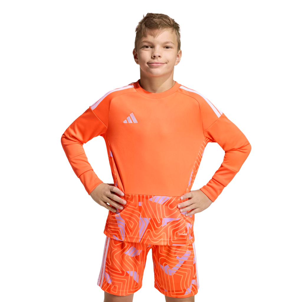 Boy in an orange long-sleeved Adidas soccer kit with a geometric pattern, posing with hands on hips against a solid black background.