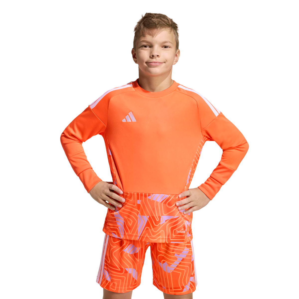 Boy in an orange long-sleeved Adidas soccer kit with a geometric pattern, posing with hands on hips against a solid black background.