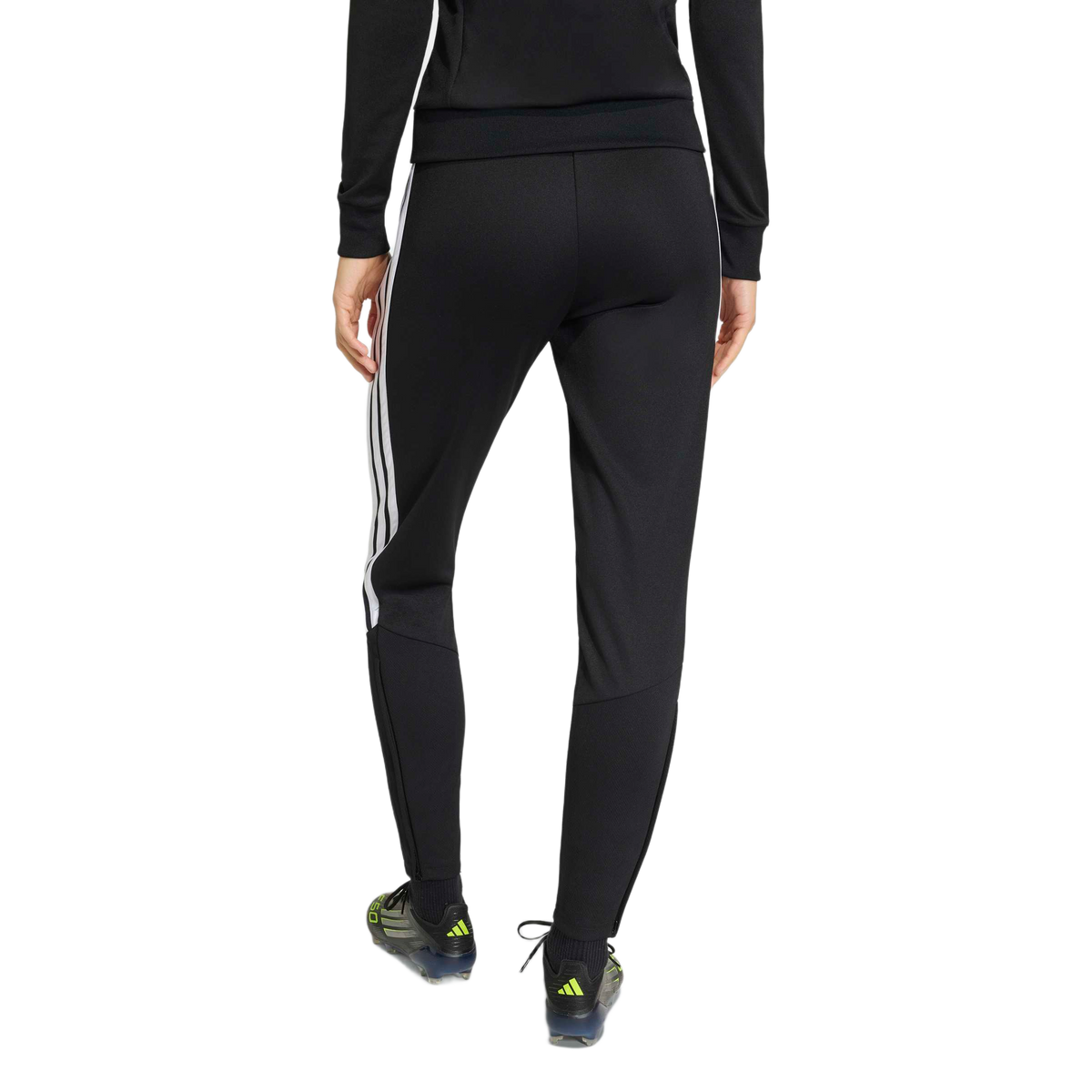 Rear view of a person wearing black Adidas track pants with white stripes on the side, a matching long-sleeved top, and soccer cleats against a light gray background.